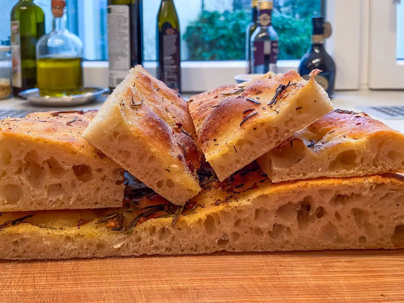 Ein leckeres Focaccia-Brot mit Kräutern, geschnitten und auf einem Holzbrett liegend. Im Hintergrund sind verschiedene Öle und Gewürze zu sehen.
