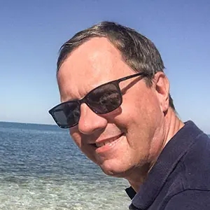 Ein lächelnder Mann mit Sonnenbrille steht vor einem Strandmeer.
